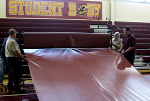 Three people installing a gym floor cover by unrolling a large protective tarp across a basketball court inside a school gym.